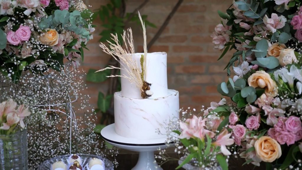 Bolo de casamento branco de dois andares sobre mesa de madeira, cercado por arranjos de flores em tons de rosa e pêssego