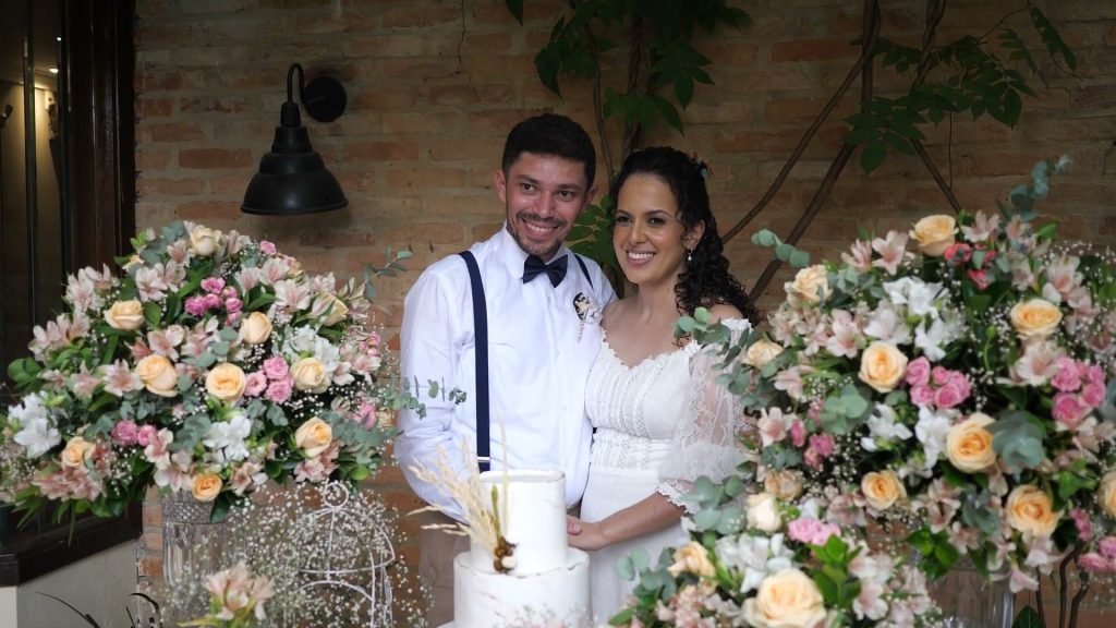Retrato dos noivos sorrindo diante da parede de flores, ao lado do bolo