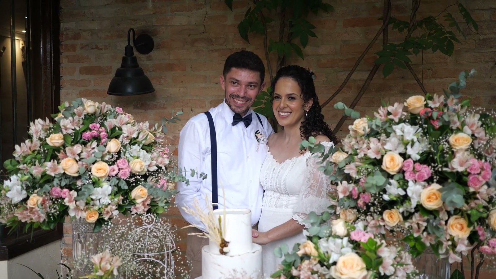 Retrato dos noivos sorrindo diante da parede de flores, ao lado do bolo