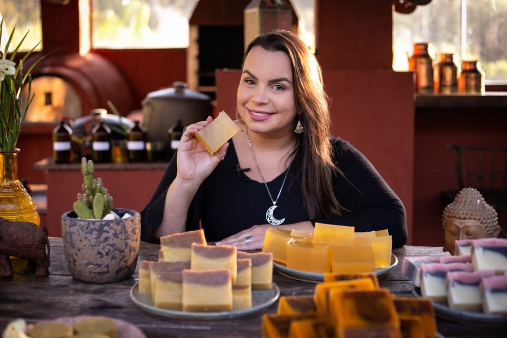 Apresentadora sentada à bancada com sabonetes artesanais coloridos à frente, sorrindo para a câmera durante a gravação do curso
