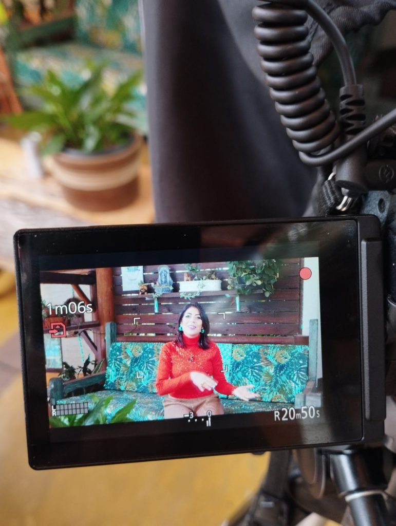 Monitor de câmera em primeiro plano mostrando a apresentadora de blusa vermelha sentada em set montado ao ar livre, entre plantas