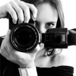 Retrato em preto e branco de Cacau Dias com a câmera diante do rosto, segurando uma Canon com flash acoplado