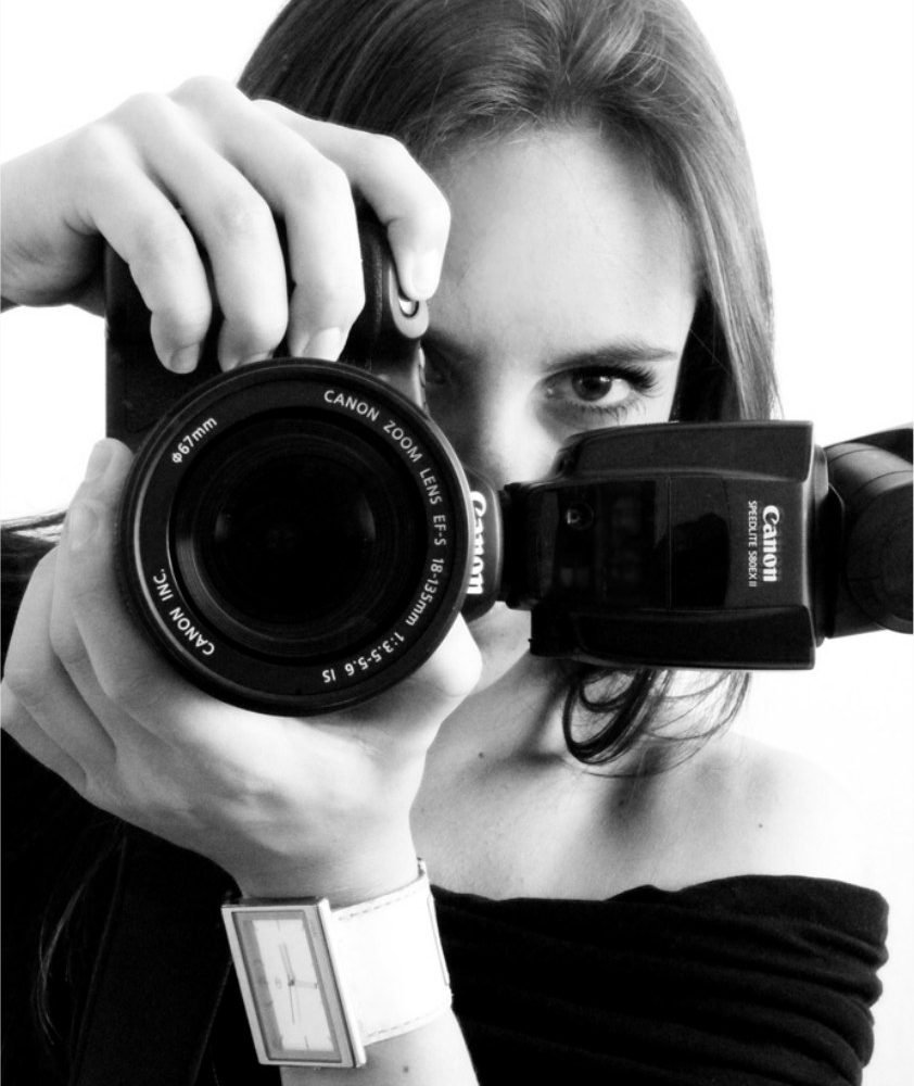 Retrato em preto e branco de Cacau Dias com a câmera diante do rosto, segurando uma Canon com flash acoplado
