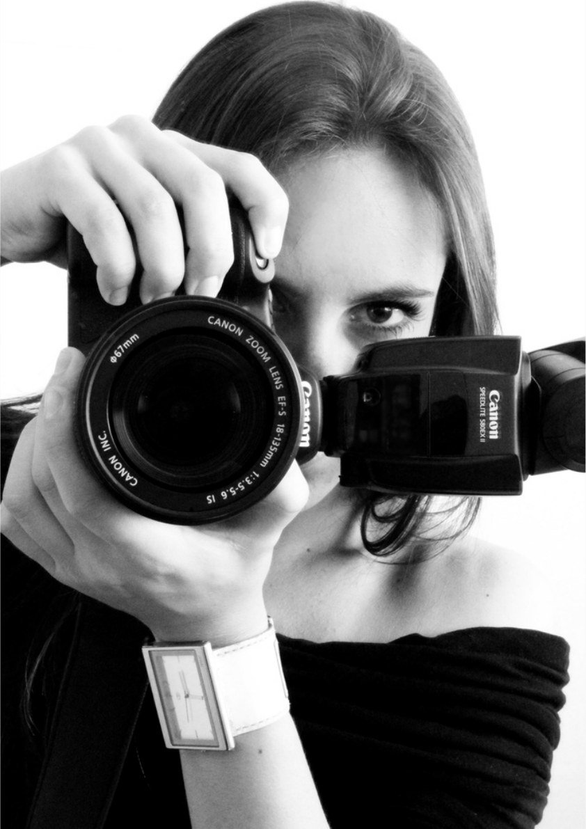 Retrato em preto e branco de Cacau Dias com a câmera diante do rosto, segurando uma Canon com flash acoplado