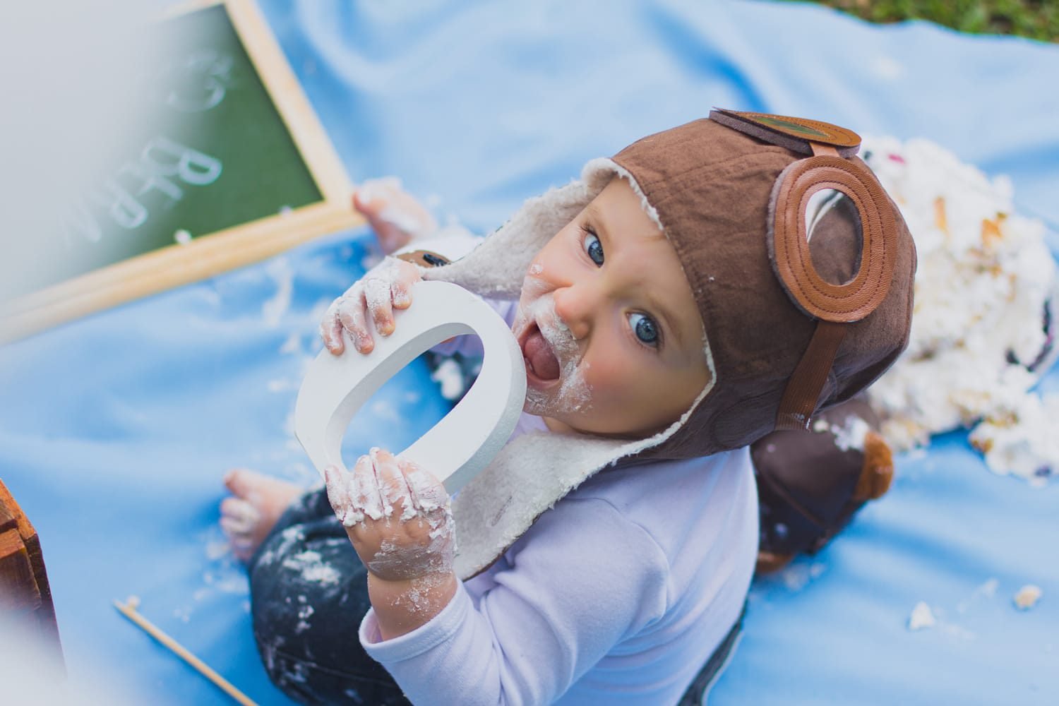 Bebê com touca de aviador sentado sobre tecido azul durante ensaio de smash the cake, coberto de chantilly, mordendo a letra O do painel de aniversário