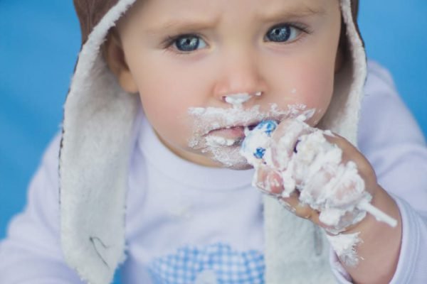 Close de bebê com touca de aviador levando as mãos cheias de chantilly à boca durante o ensaio de smash the cake ao ar livre