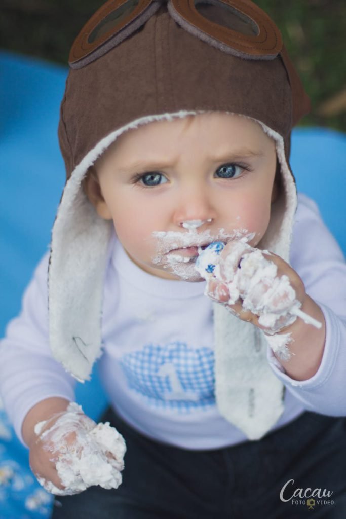 Close de bebê com touca de aviador levando as mãos cheias de chantilly à boca durante o ensaio de smash the cake ao ar livre